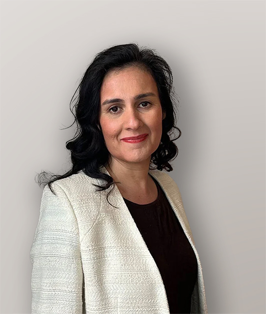 A woman with long, wavy black hair, wearing a white textured blazer over a dark top, smiles softly while standing against a plain light gray background.