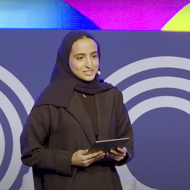 A woman wearing a black hijab and dark clothing stands on stage holding a tablet, speaking in front of a blue background with white circular patterns and colorful lights above.