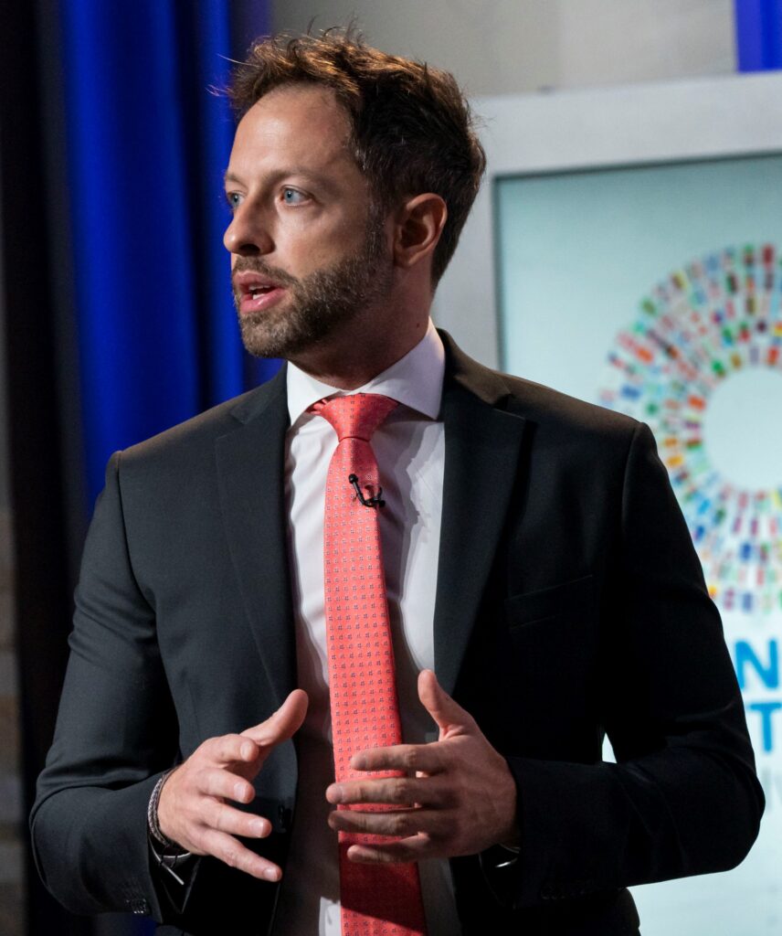 A man with short brown hair and a beard, wearing a black suit, white shirt, and red tie, speaks while gesturing with his hands. Behind him is a screen displaying a colorful circular graphic.