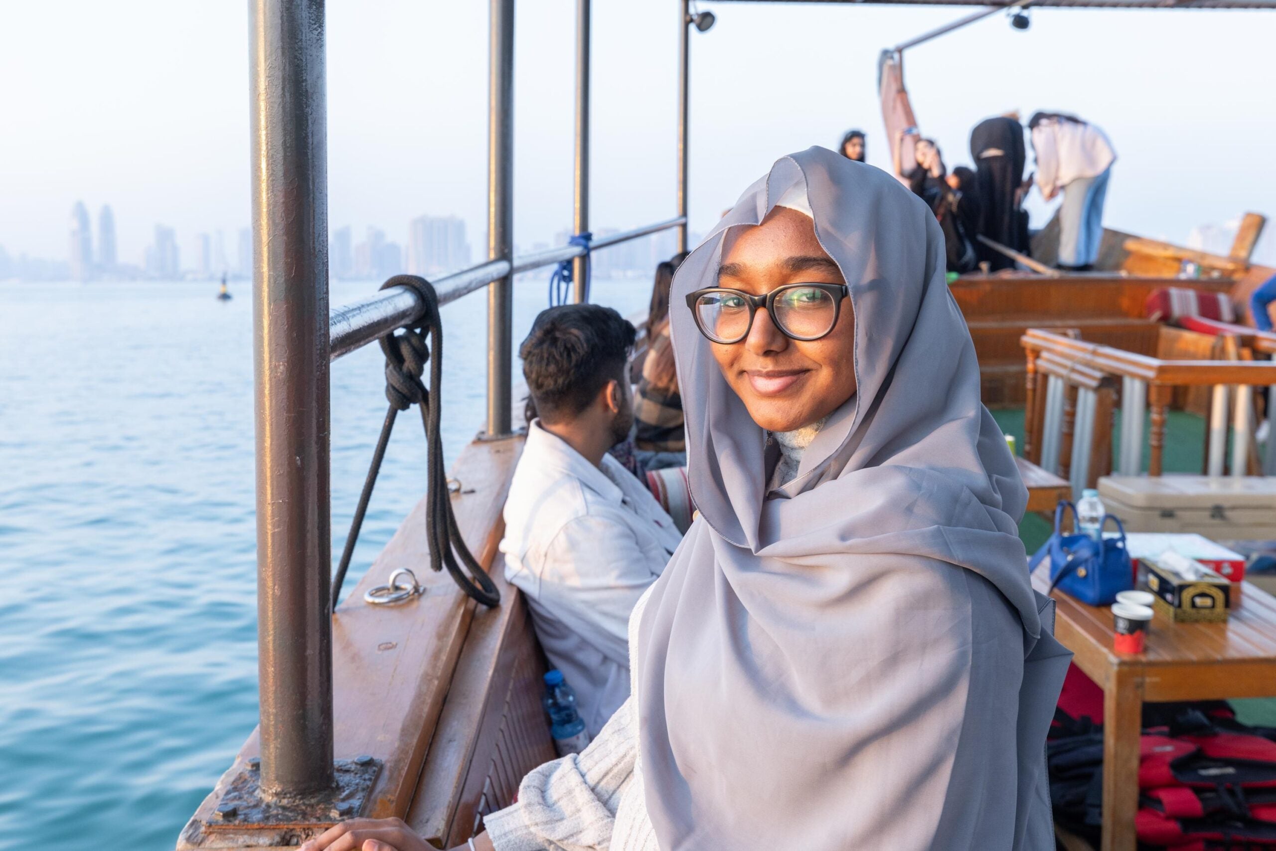 A woman in a gray hijab and glasses smiles while sitting on a wooden boat with other passengers; city buildings and water are visible in the background.