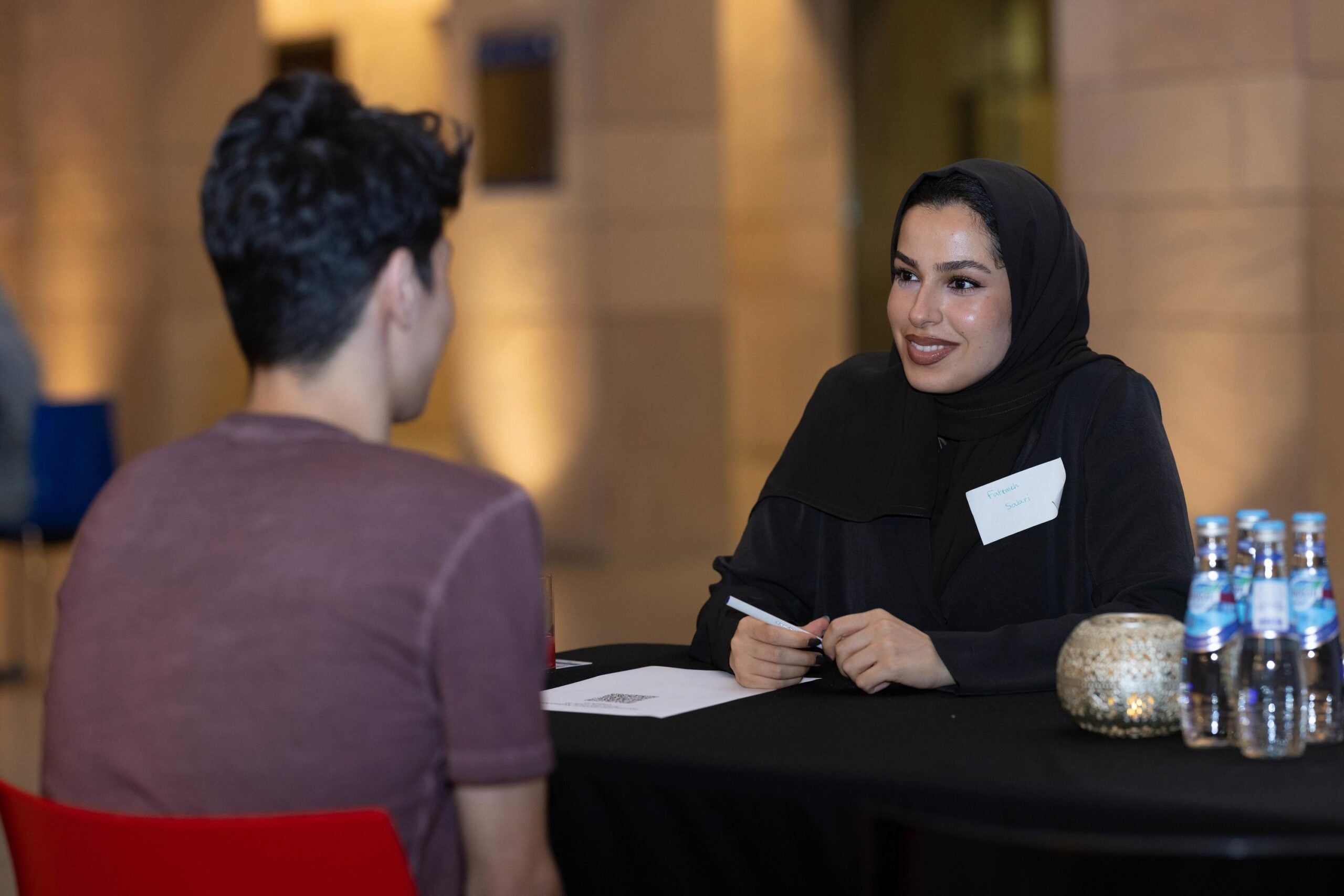 A woman in a black hijab and abaya, wearing a name tag, smiles while talking to a man in a maroon t-shirt across a table with water bottles and a decorative item on it. The background is softly lit.