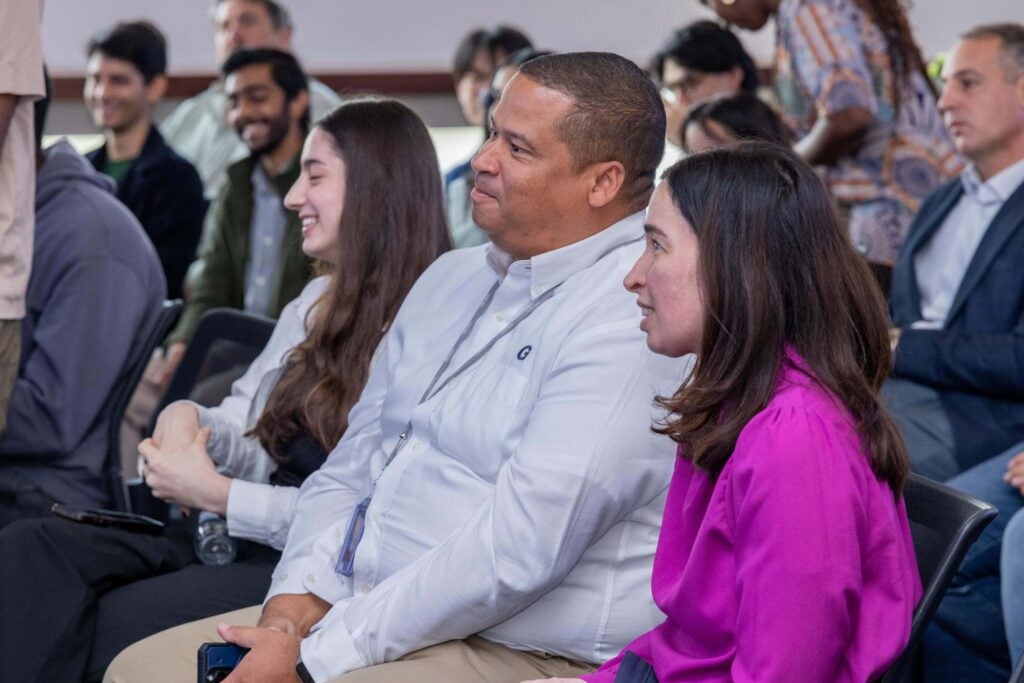 A group of people sit in rows at an indoor event, listening attentively. Three individuals in the front—two women and one man—are focused on something off-camera, while others are seated behind them.