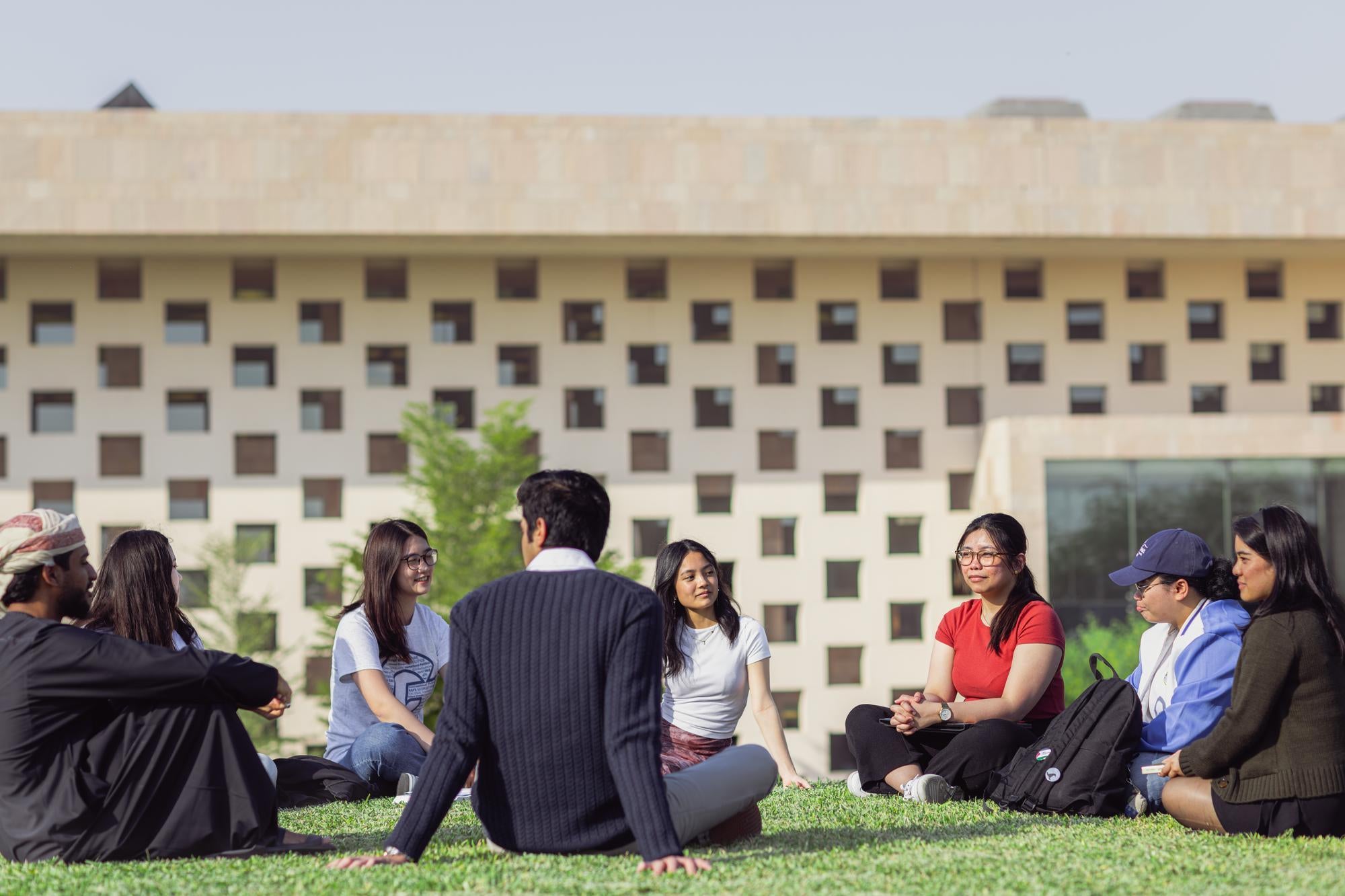 A group of students sits in a circle on grass, talking and smiling, with a modern building featuring square windows in the background on a sunny day.