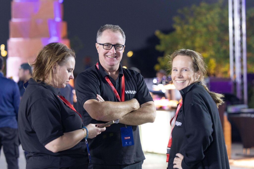 Three people wearing black shirts and red lanyards stand outdoors at night. One looks at a phone, one stands with arms crossed smiling, and one smiles at the camera. Background is blurred with lights and trees.