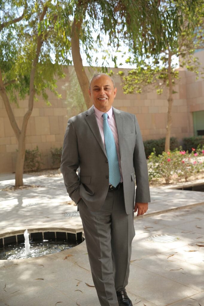 A man in a gray suit, pink shirt, and blue tie stands outdoors on a sunny day, smiling with hands in his pockets. He is the dean Safwan Masri.