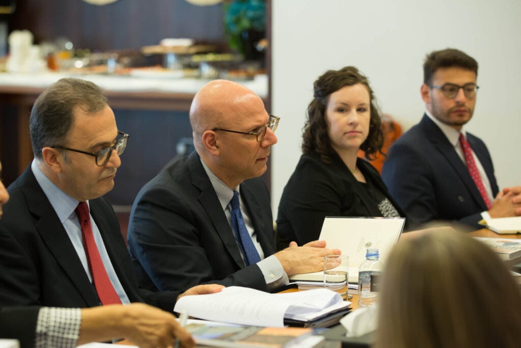 A group of professionals sit around a conference table engaged in discussion. Documents, notebooks, and drinks are on the table. The focus is on a man in glasses speaking, with others listening attentively.