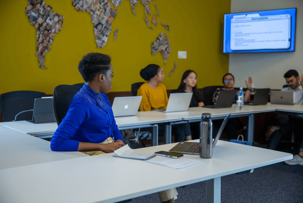 A group of people sit around a conference table with laptops, engaged in discussion. One person in the foreground looks toward others. A wall display screen shows text, and a collage of faces decorates the wall.