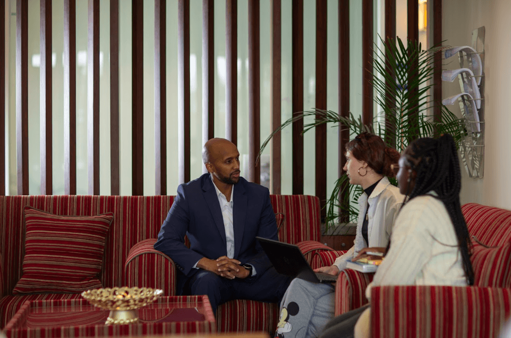 Three people sit on red striped couches in a modern office lounge, having a discussion. One man in a suit faces two women, one with a laptop, with vertical wooden slats and plants in the background.