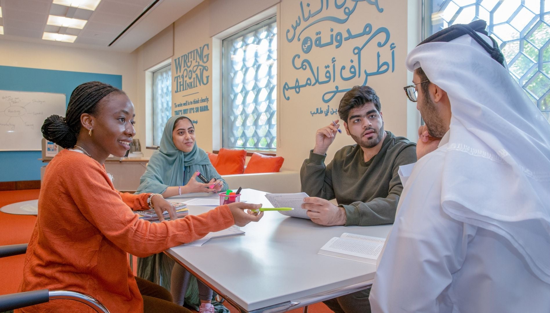 Four students sit around a table in a bright study room, discussing and taking notes. The room has colorful walls, Arabic and English writing, and large windows letting in natural light.