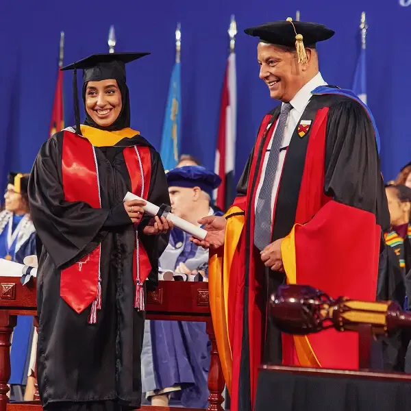 A smiling graduate in a cap and gown receives a diploma from a faculty member in academic regalia during a graduation ceremony, with flags and other graduates visible in the background.