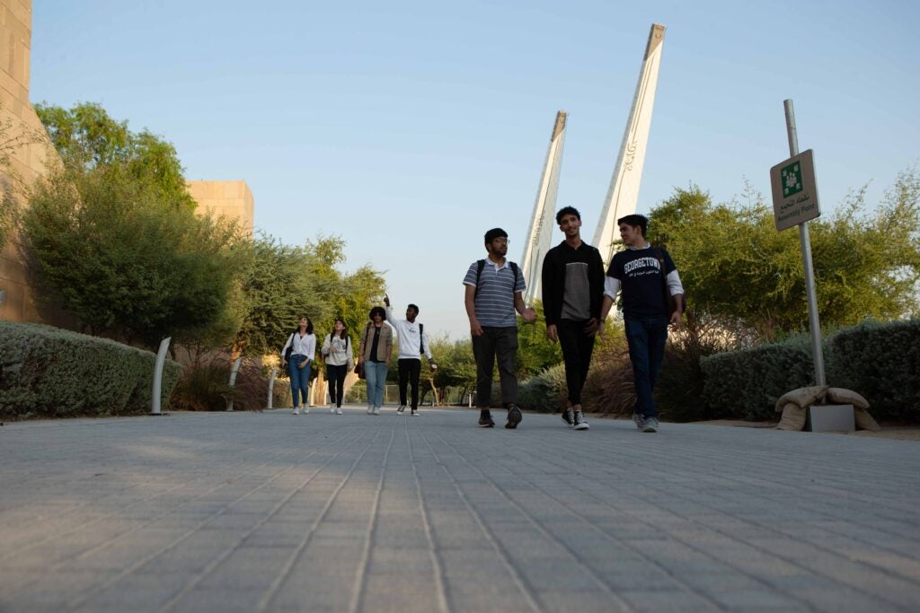 A group of students walks along a tree-lined campus pathway on a sunny day, some engaged in conversation, with modern buildings and tall structures in the background.