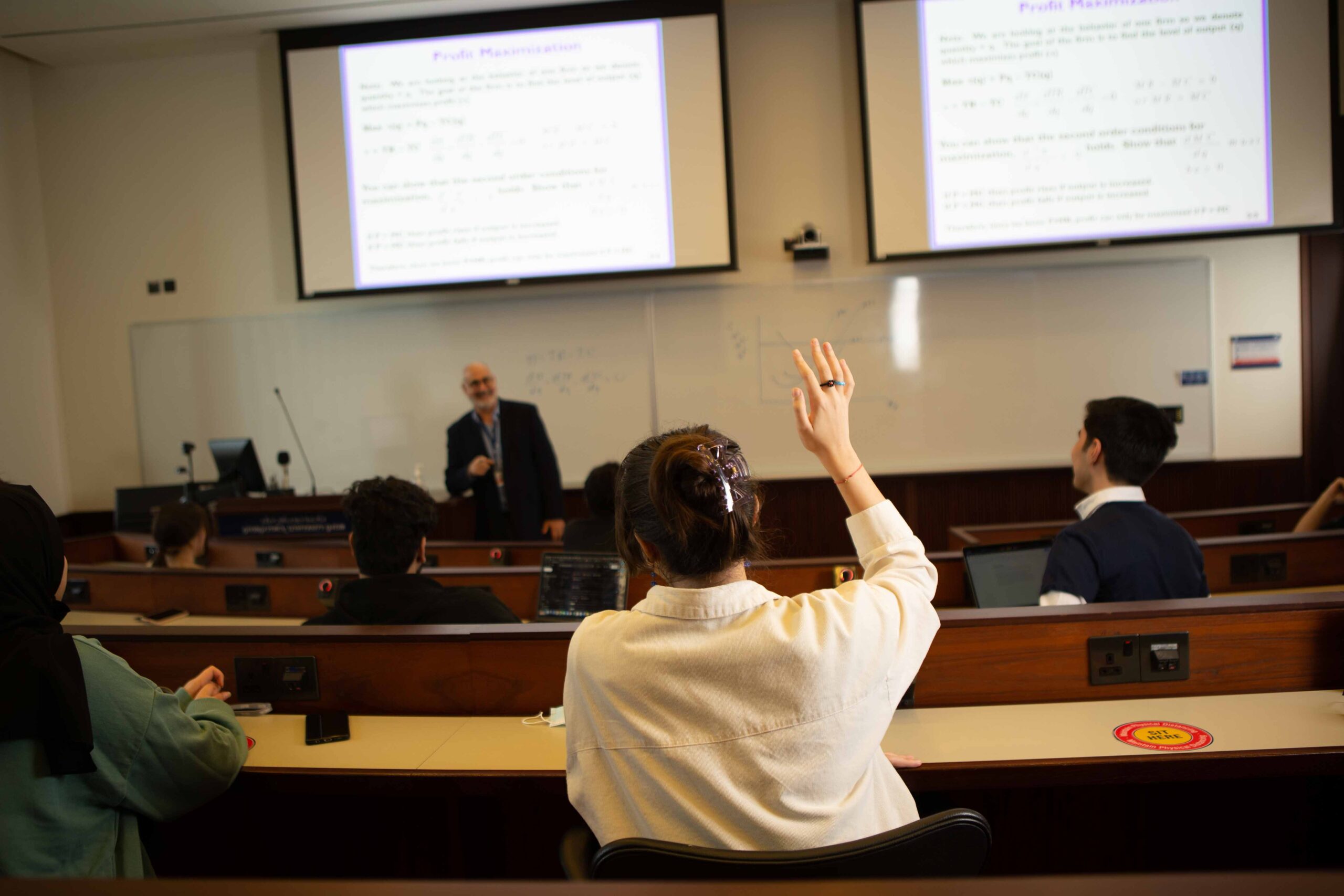 A student raises their hand in a university classroom while a professor lectures at the front. Several students are seated at desks, and two large screens display slides titled Profit Maximization.