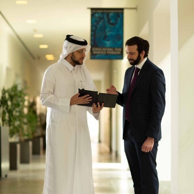 Two men stand in a hallway, one wearing traditional Middle Eastern attire and the other in a business suit, looking at a tablet together and engaged in conversation.