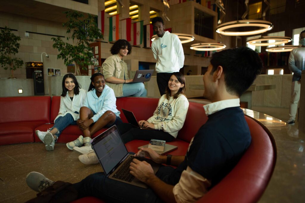 A group of six diverse students sits on red couches in a modern building, smiling and working on laptops, with international flags hanging in the background.