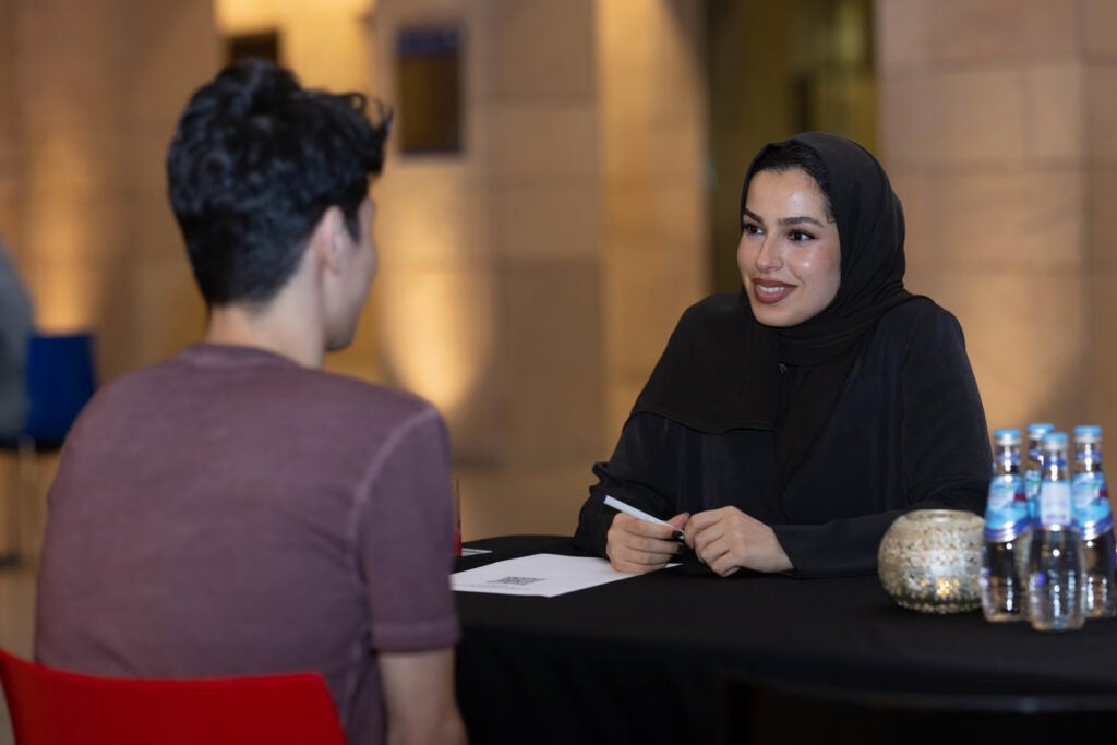 A woman in a black hijab sits at a table, smiling and speaking to a man with short dark hair. Several water bottles and a decorative candle are on the table, with a warmly lit background.