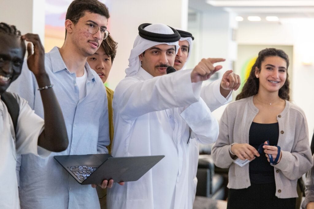 A man in traditional Middle Eastern attire gestures and points while standing with a group of diverse young adults, one holding a laptop. They appear engaged in conversation, smiling in a bright indoor setting.