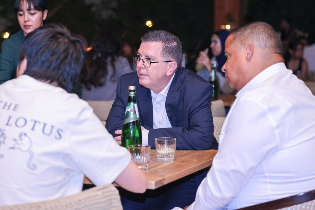 Three people sit at a wooden table in conversation at an event. One man in a suit and glasses faces two others. Bottles of sparkling water and glasses are on the table; other guests are blurred in the background.