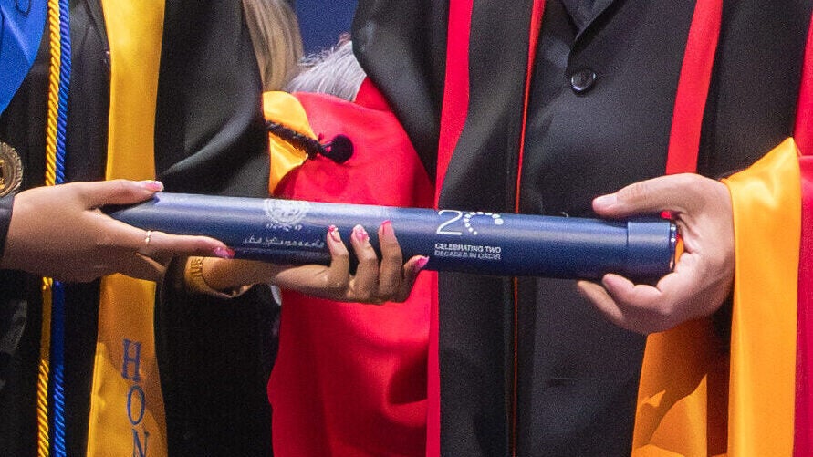 A graduate in a cap and gown with honor cords and sashes receives a diploma from a faculty member in academic regalia during a graduation ceremony.