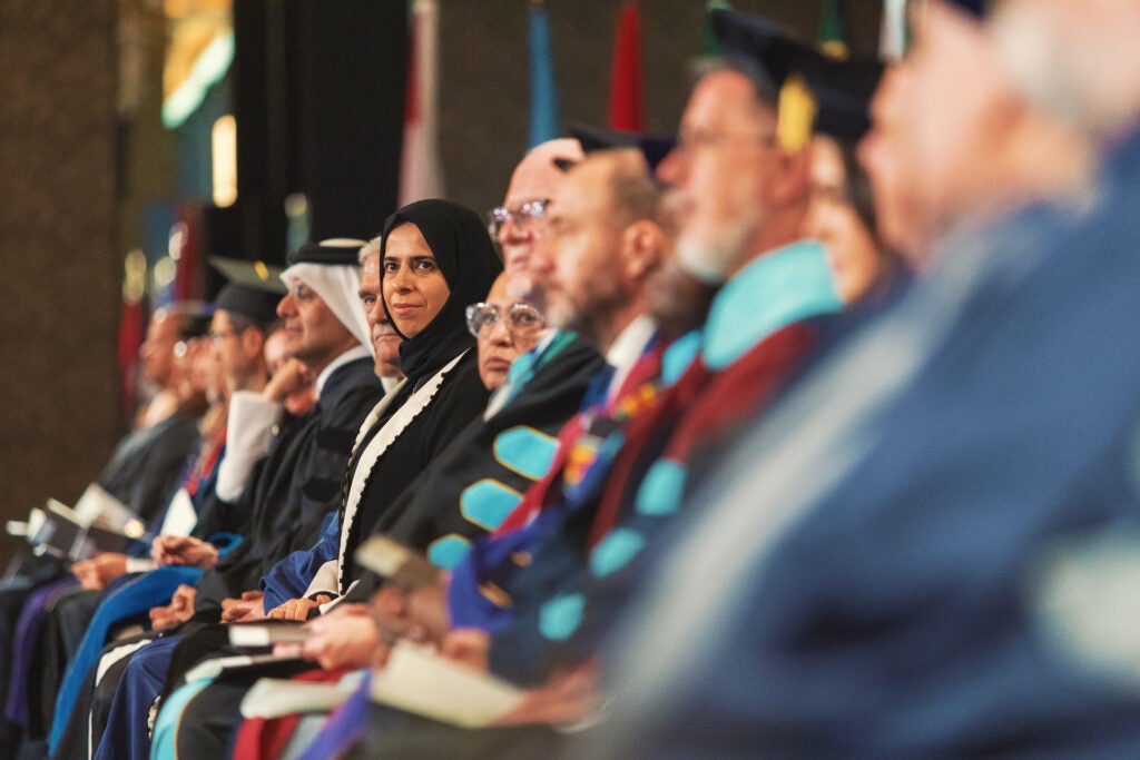 A group of faculty members in academic regalia sit in a row at a graduation ceremony, with one woman in a black hijab and gown looking directly at the camera among colleagues.