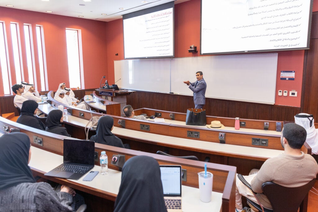 Teacher stands at the head of a classroom with tiered seats. Students with open computers look at the teacher. On the screens behind the teacher is Arabic text