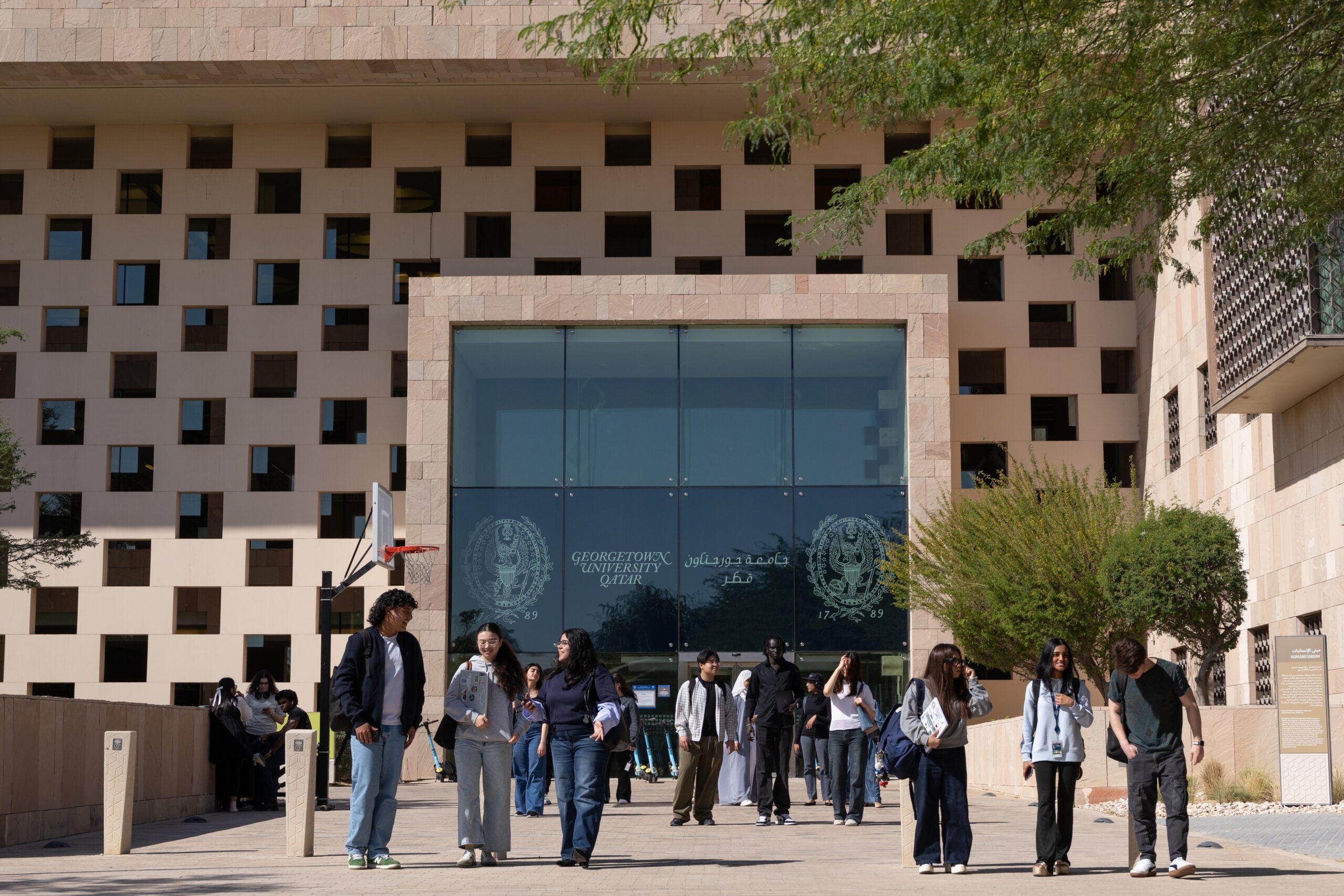 Students walk and talk in front of a modern university building with a patterned facade and large glass entrance at Georgetown University in Qatar. Trees and groups of people are visible in the background.
