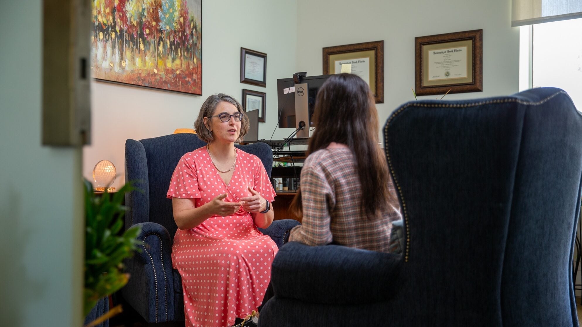 A woman in a pink polka-dot dress speaks with another woman in an office furnished with blue chairs, wall art, and framed certificates. The scene appears to depict a counseling or therapy session.
