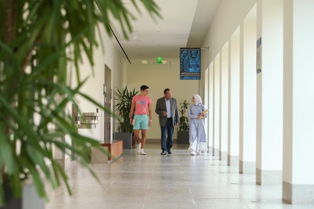 Three people, two men and one woman in a white headscarf, walk and talk in a bright indoor hallway with large windows and green plants.
