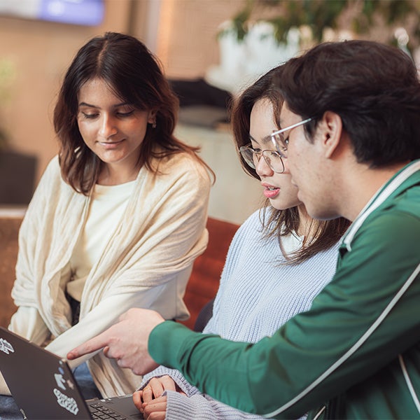 Three young adults sit together, focused on a laptop. One person points at the screen while the others watch attentively, appearing engaged in a collaborative discussion or study session.