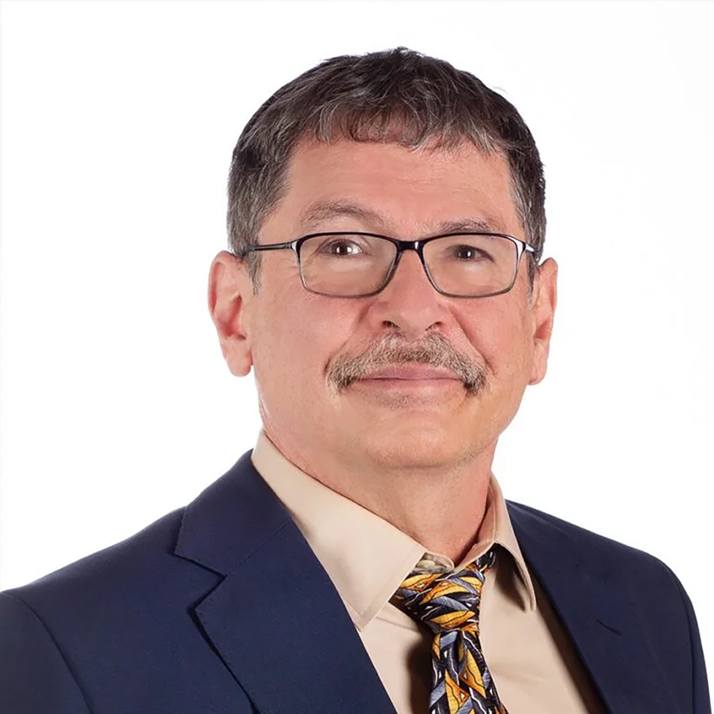 A middle-aged man with short brown hair, glasses, and a mustache, wearing a navy suit, beige shirt, and patterned tie, smiling slightly against a plain white background.