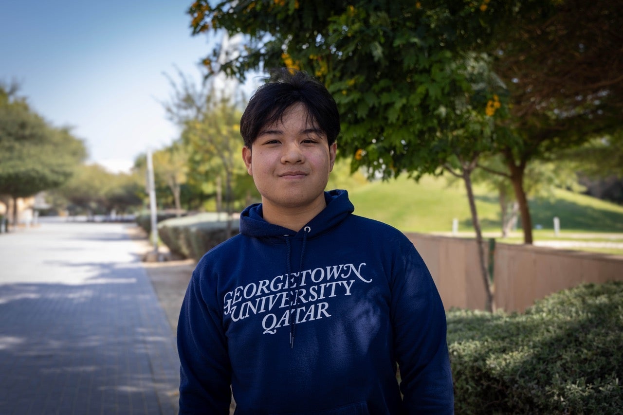 A person wearing a Georgetown University Qatar hoodie stands outside on a sunny day, with trees, greenery, and a pathway in the background.