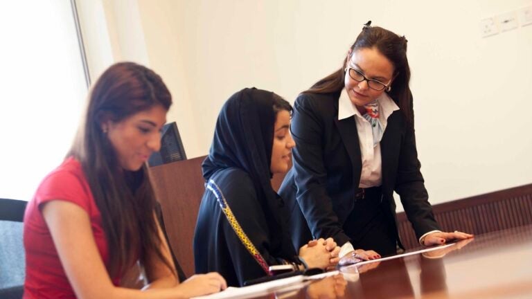 Three women in an office setting are working together at a desk. Two are seated, one wearing a hijab and the other in a red shirt, while a woman in a suit stands, leaning over to discuss papers with them.