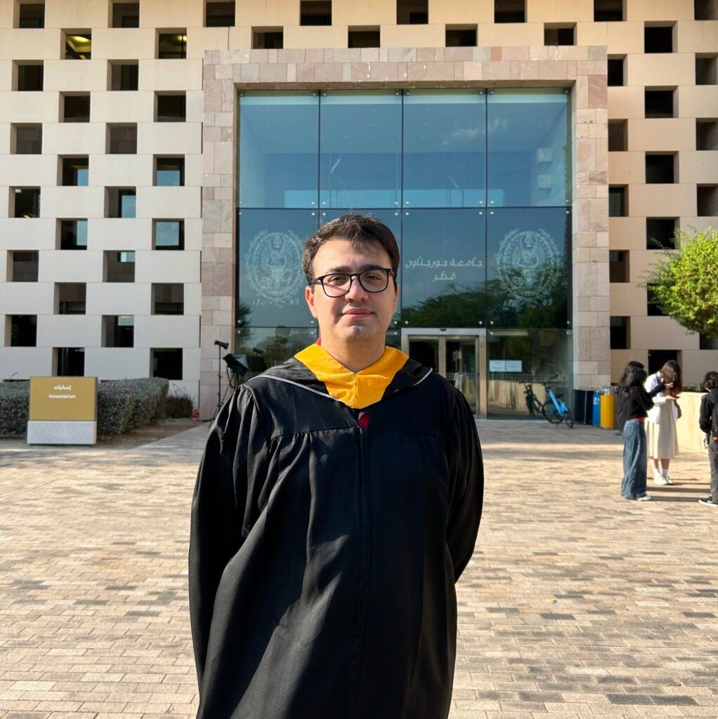 Aras Karlidag wearing a graduation gown and hood stands outside a modern building with large windows and a stone facade, and GU-Q branding with Arabic Writing. Other people are in the background, and sunlight casts shadows on the paved ground.