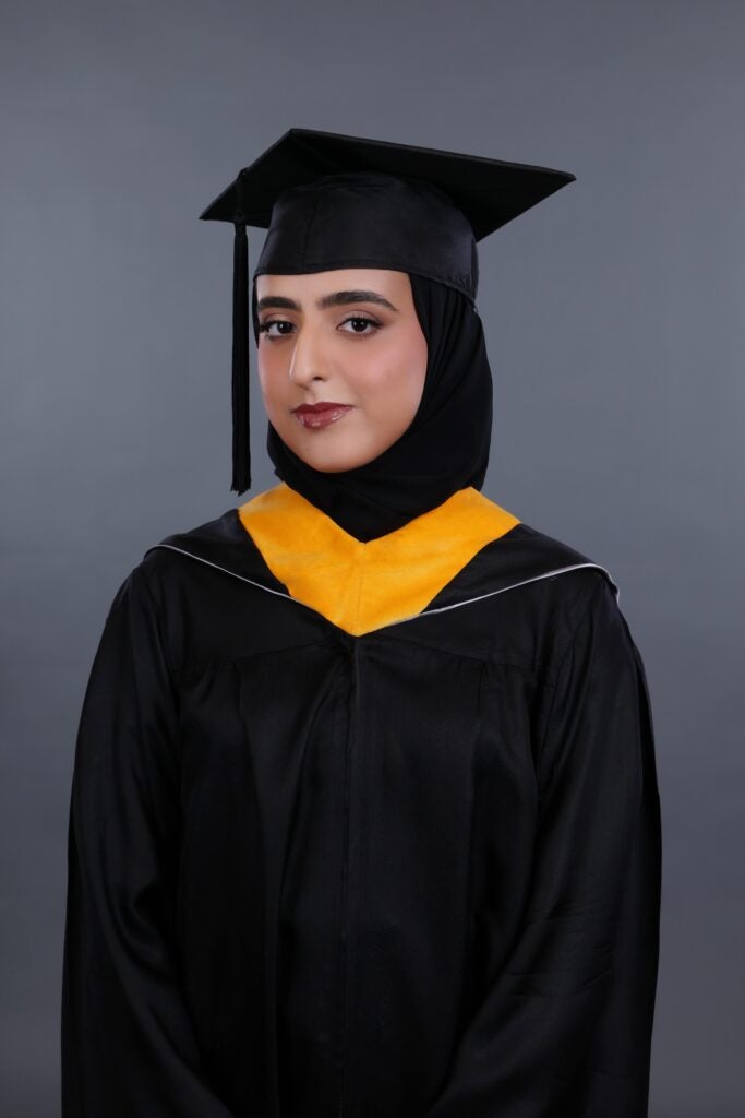 A woman wearing a black graduation gown and cap with a yellow stole smiles slightly, posing in front of a plain gray background. She is also wearing a black hijab under her cap.