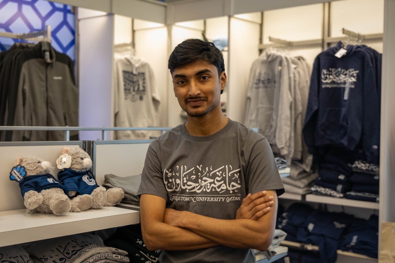 A young man stands with arms crossed in a campus store, surrounded by university-branded hoodies and two stuffed animals on a shelf.