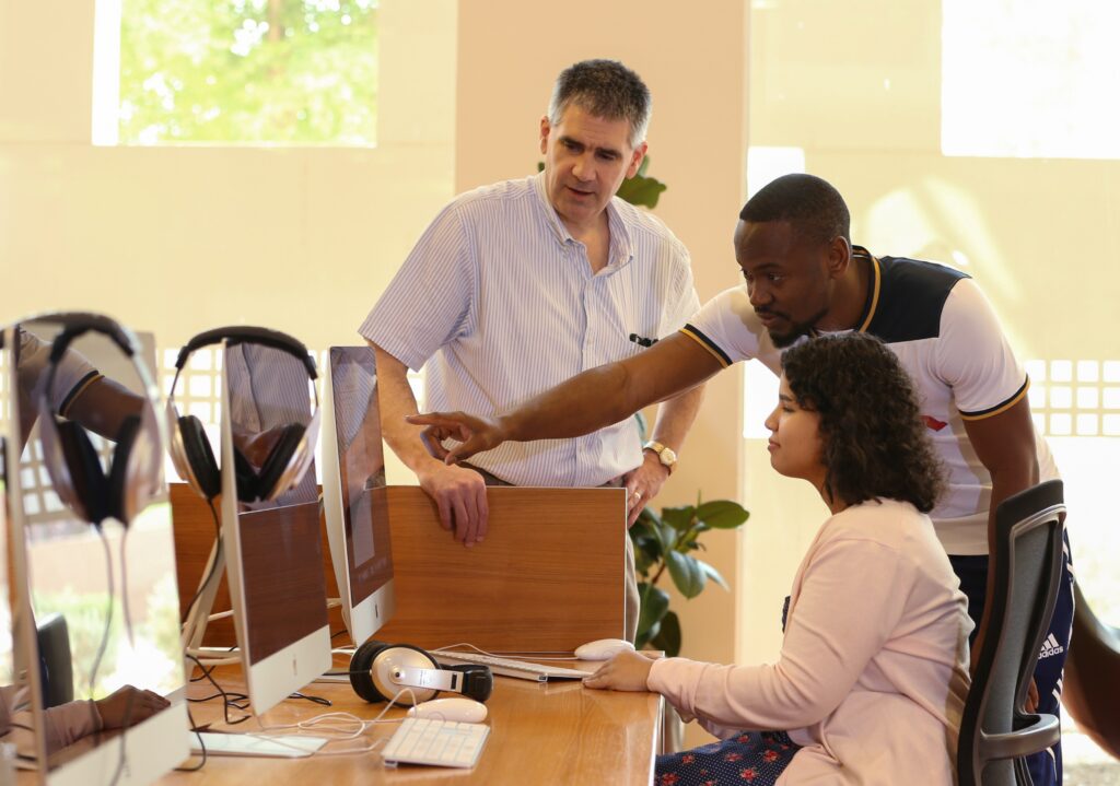 Three people in a computer lab: one man standing, another man leaning in and pointing at a computer screen, and a seated woman using the computer, all appearing to collaborate on a task.