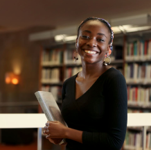 A young woman wearing a black top smiles while holding a book in a library, with shelves of books visible in the background.
