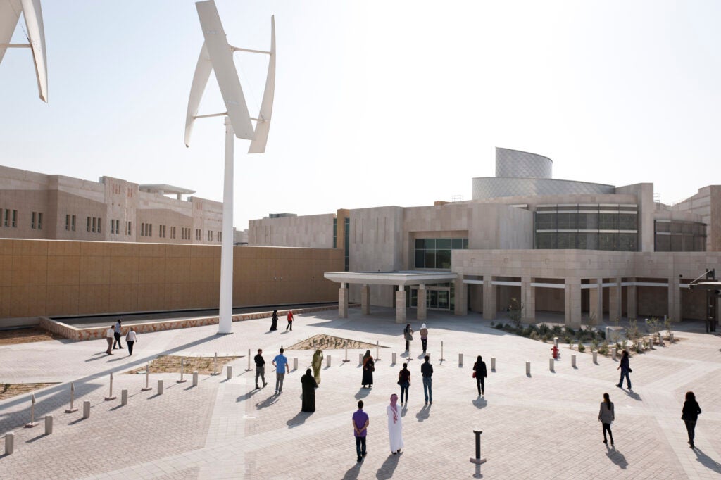 People walk across a courtyard with modern wind turbines in front of a large, contemporary building under a bright sky.