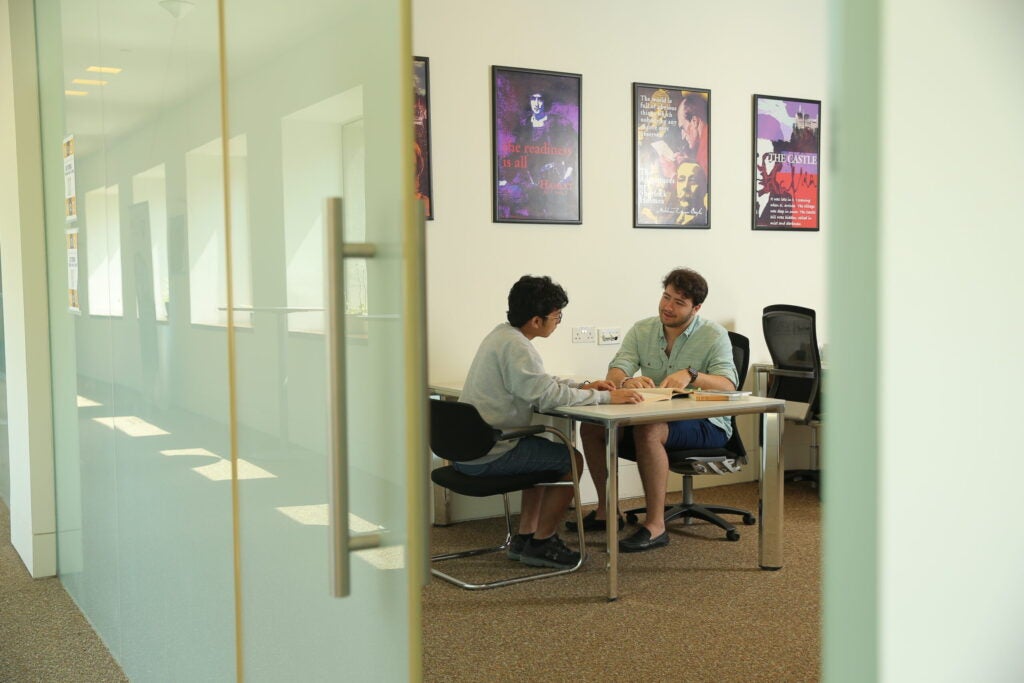 Two people sit across from each other at a desk in a bright room, engaged in conversation or study. Books are open on the desk, and colorful posters hang on the wall behind them.
