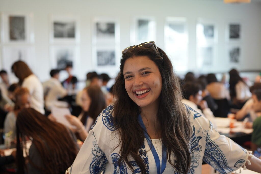 A young woman with long dark hair and a big smile stands in the foreground of a busy, brightly lit room filled with people working or studying at tables. She wears a white blouse with blue patterns and sunglasses on her head.