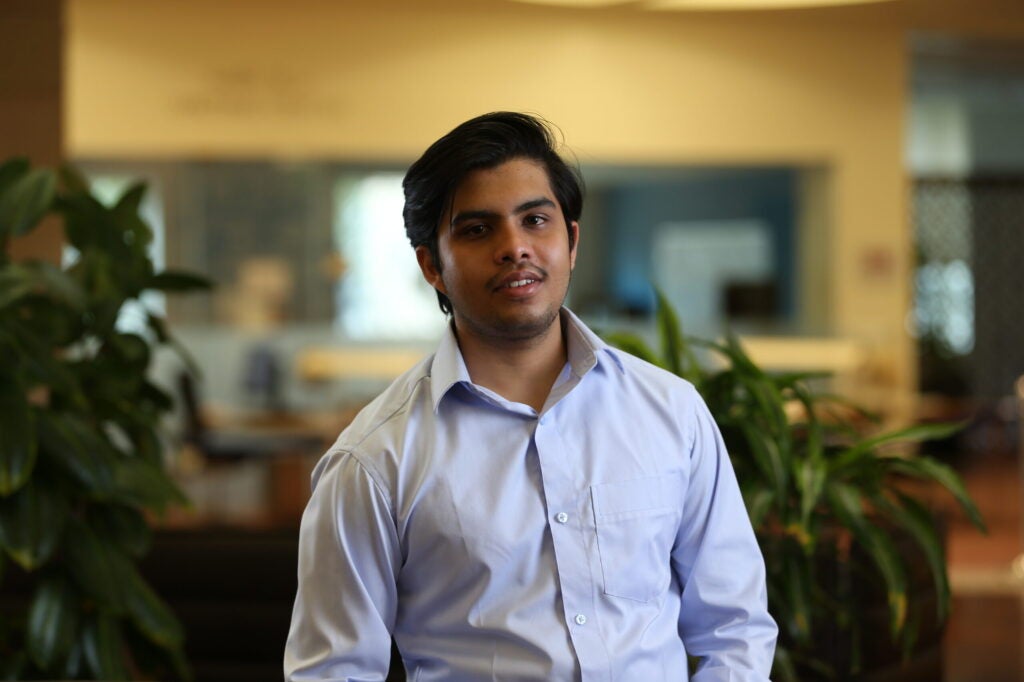 A young man with dark hair, wearing a light blue button-up shirt, stands indoors in a well-lit room with plants and blurred office furnishings in the background.