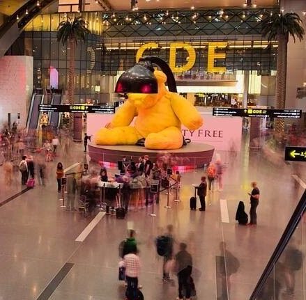 A large yellow teddy bear sculpture with a lamp over its head stands in a busy airport terminal with people walking around and signs for gates visible.