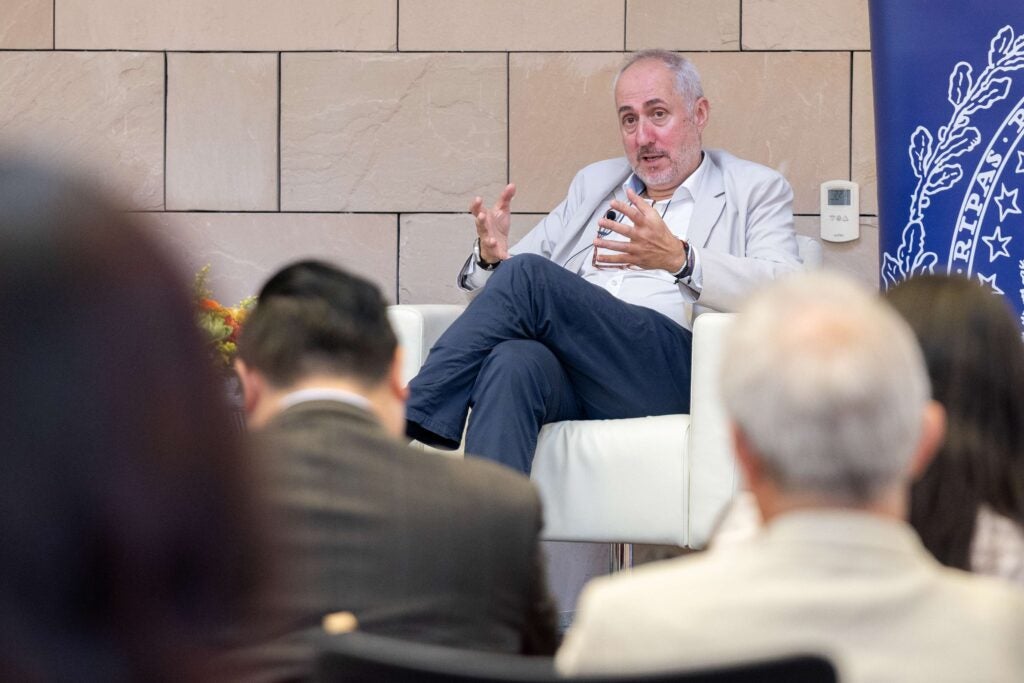 A man in a light-colored suit speaks and gestures while seated on a white chair during a panel discussion. He addresses an audience, some of whom are visible in the foreground.