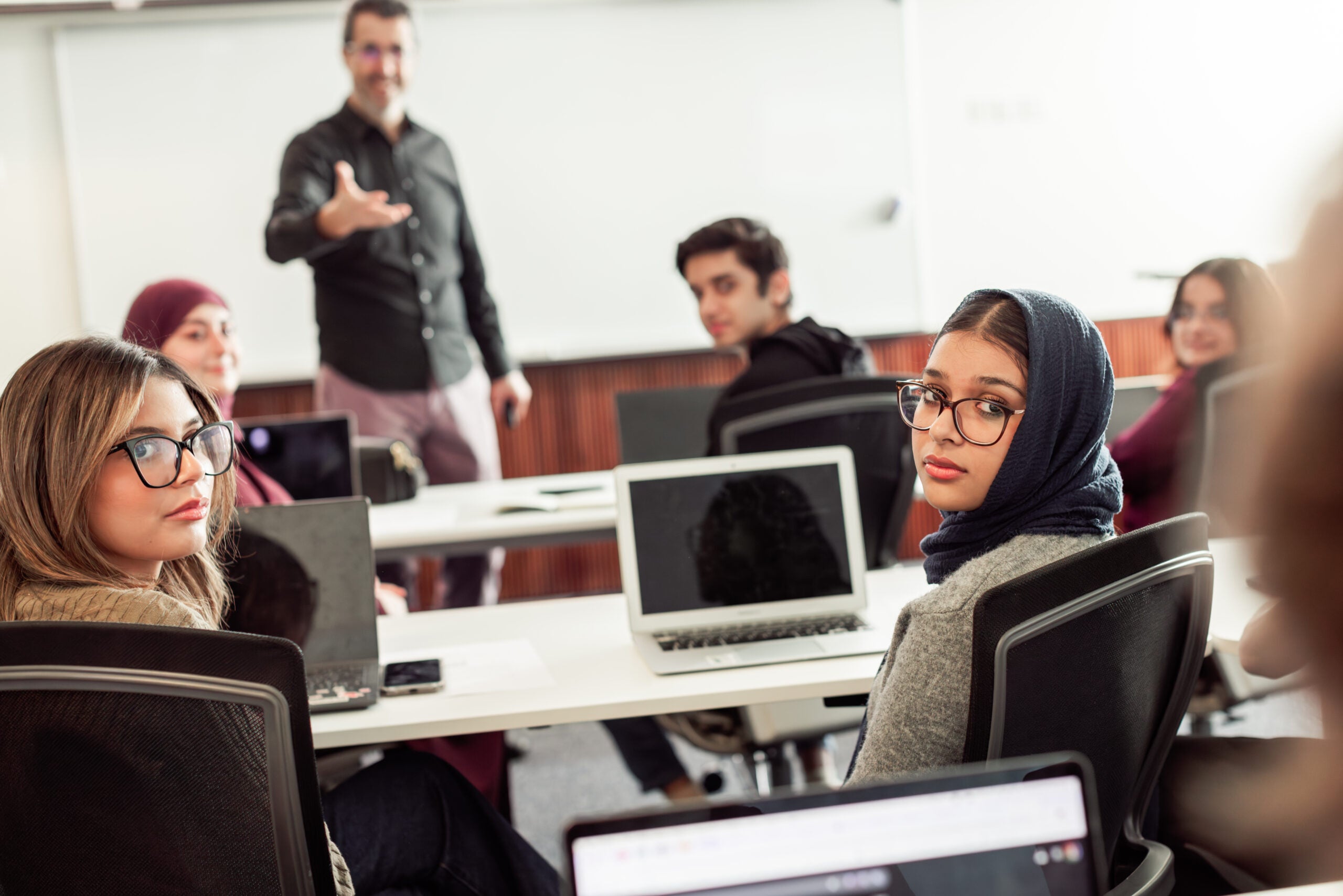 A group of students sitting at desks with laptops, turning to look at a teacher standing and gesturing at the front of a classroom with a whiteboard in the background.