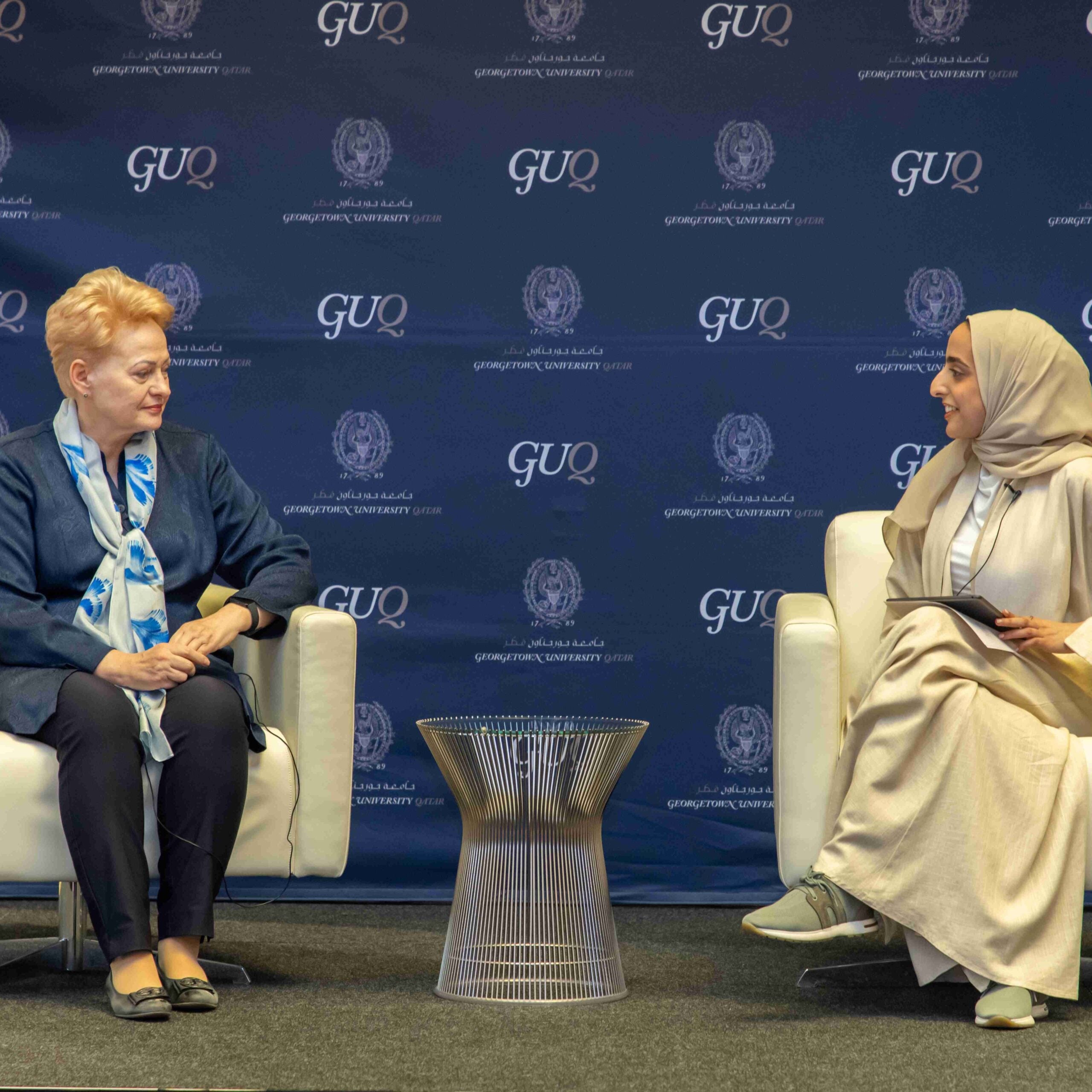Two women sit facing each other on stage in white chairs, engaged in conversation. Behind them is a Georgetown University in Qatar backdrop and a small table is placed between them. One woman holds a notepad.