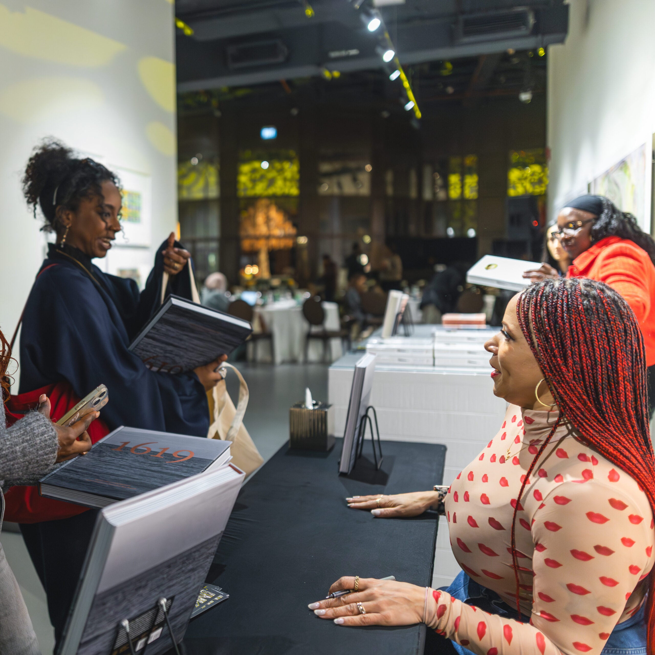 Two women stand at a table holding books, smiling and interacting with a seated woman behind the table at a book signing event. Other attendees are visible in the background.