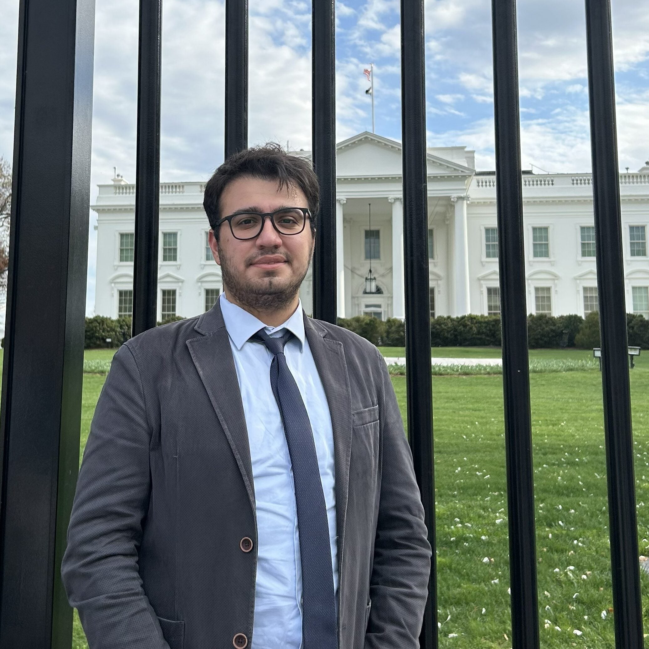 A man in a suit and tie stands in front of a black metal fence with the White House and green lawn visible in the background under a partly cloudy sky.