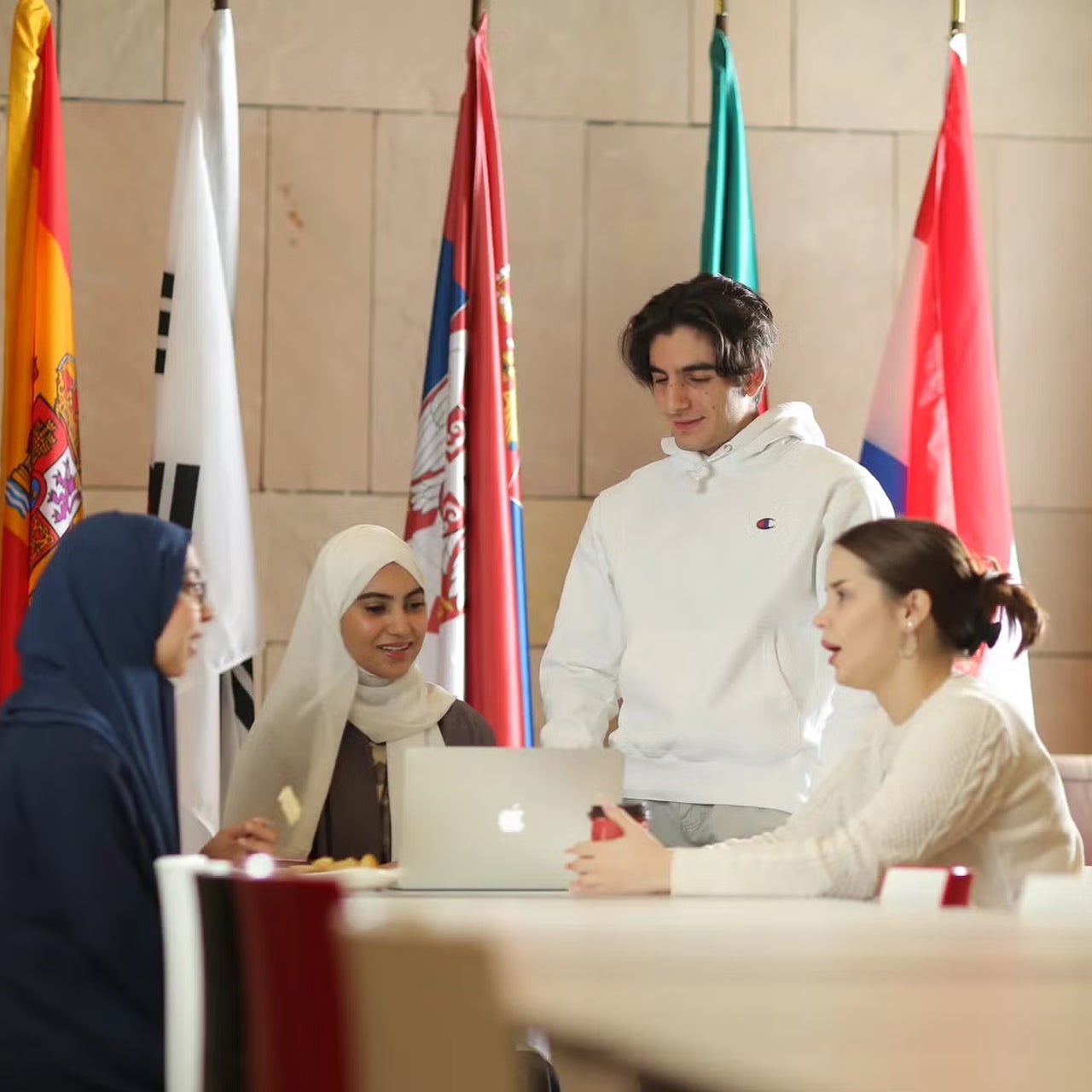 Four young adults, three women and one man, sit and stand around a laptop having a discussion. International flags are displayed in the background. The setting appears to be a study or meeting area.
