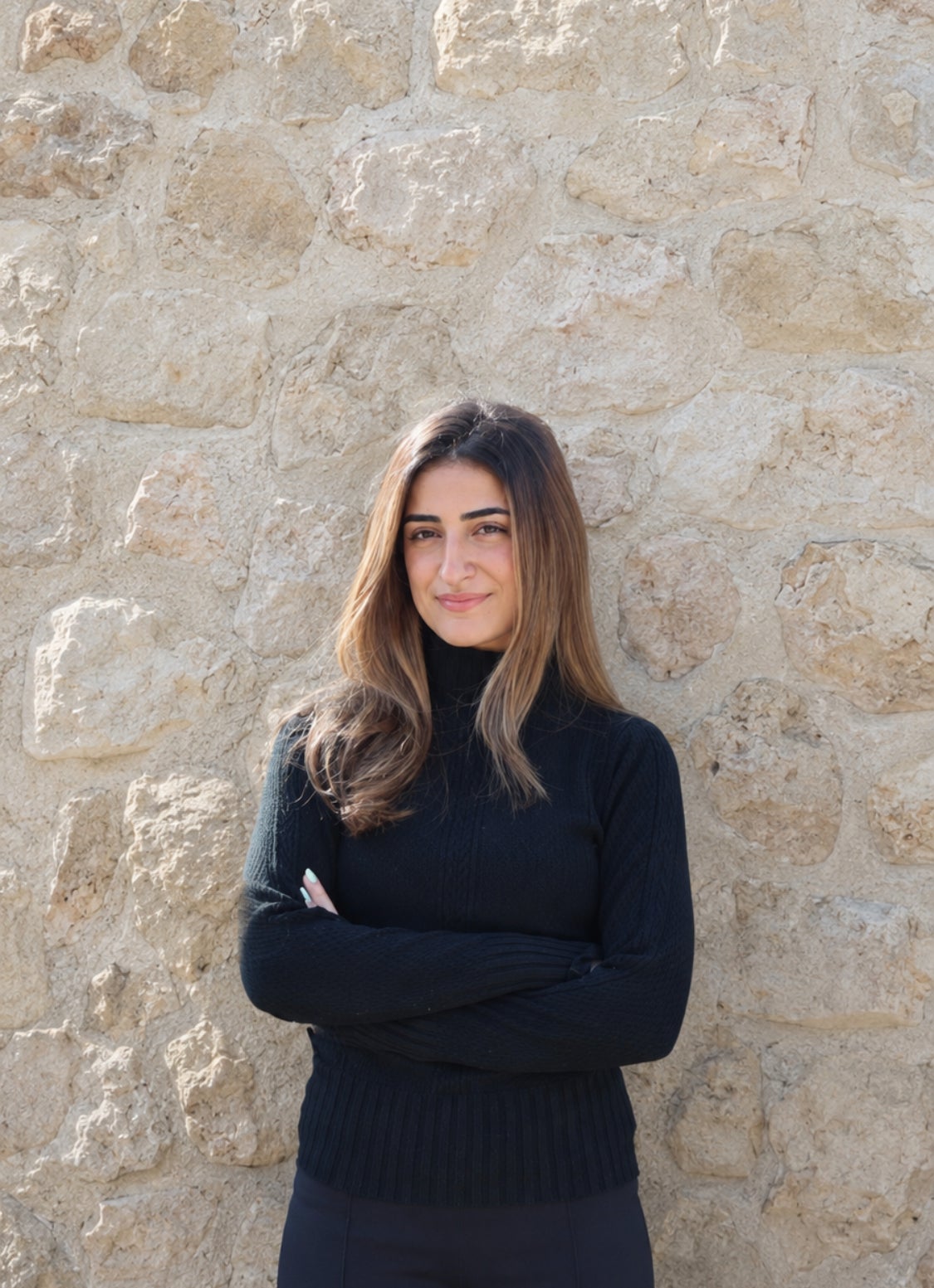 A woman with long brown hair, wearing a black turtleneck sweater, stands with arms crossed in front of a textured stone wall, looking at the camera and smiling slightly.