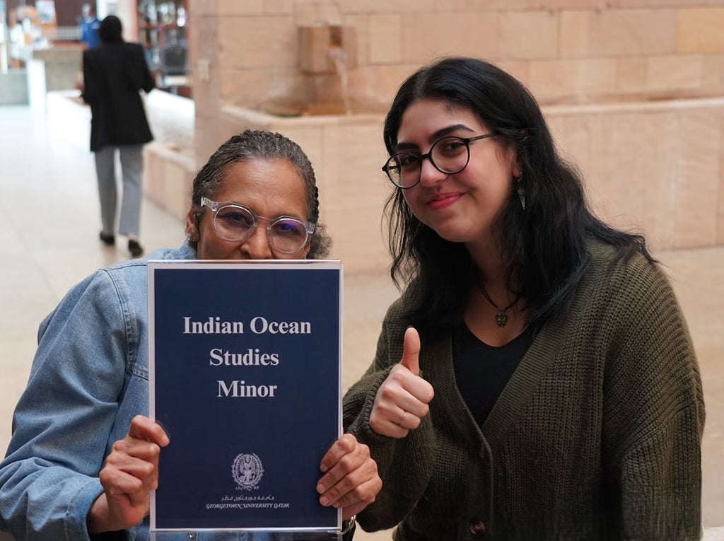 Two people smile at the camera; one holds a sign reading Indian Ocean Studies Minor while the other gives a thumbs up. They appear to be in a spacious indoor area with tan walls.