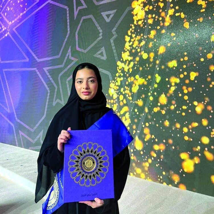 A woman in a black hijab holds a blue folder with a gold design, standing in front of a brightly lit, geometric-patterned backdrop with golden sparkling lights.
